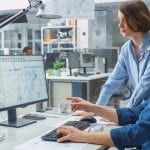 Engineer Talking With Project Manager and Working on Desktop Computer Using CAD Software with Technical Drawings on the Screen. In the Background Engineering Facility Specialising on Industrial Design