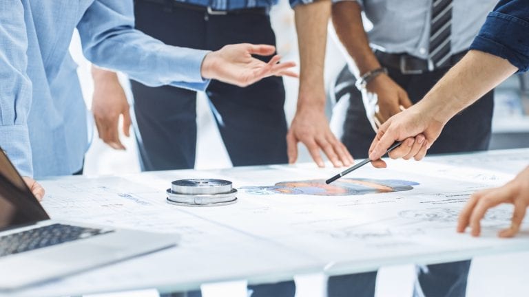 Industrial Engineering Facility: Close-up on Hands of Group of Engineers, Technicians and Specialists on a Meeting, Tracing Lines and Analysing Engine Design Technical Drafts Laying on the Table