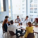 Young colleagues, caucasian business partners working as designers sit together at table in modern office with panoramic window. Papers, colorful pencils on table, active discussion