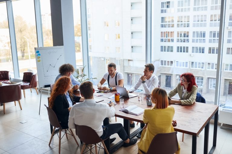 Young colleagues, caucasian business partners working as designers sit together at table in modern office with panoramic window. Papers, colorful pencils on table, active discussion